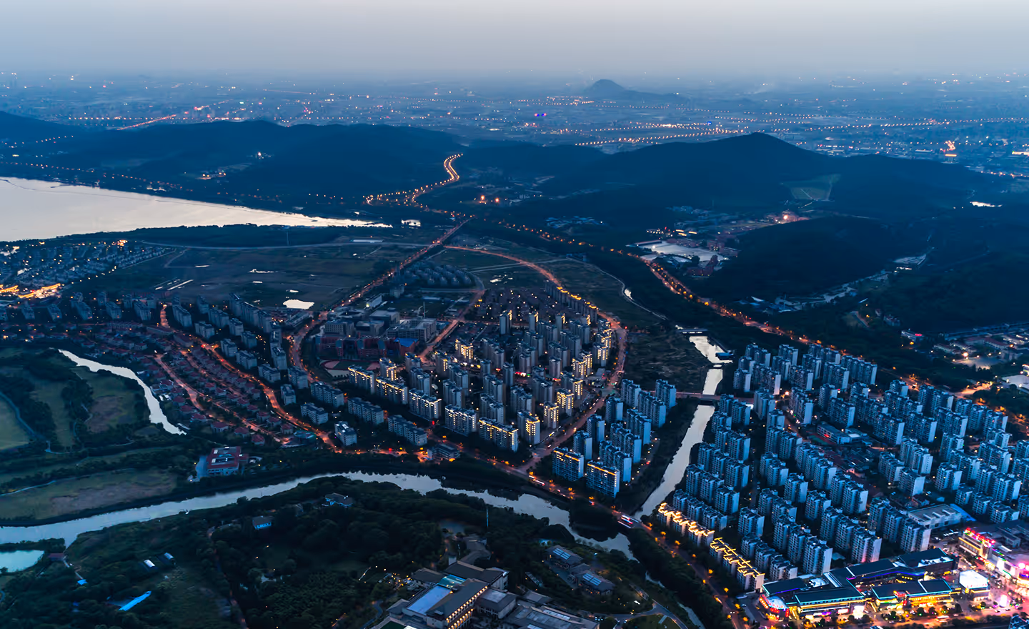 Aerial evening view of a riverside city with illuminated buildings, winding roads, and distant hills under a dim sky.
