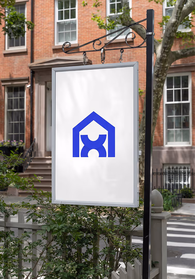 White hanging sign with a blue house-shaped logo featuring two stylized figures inside, set against a background of brick buildings and greenery.